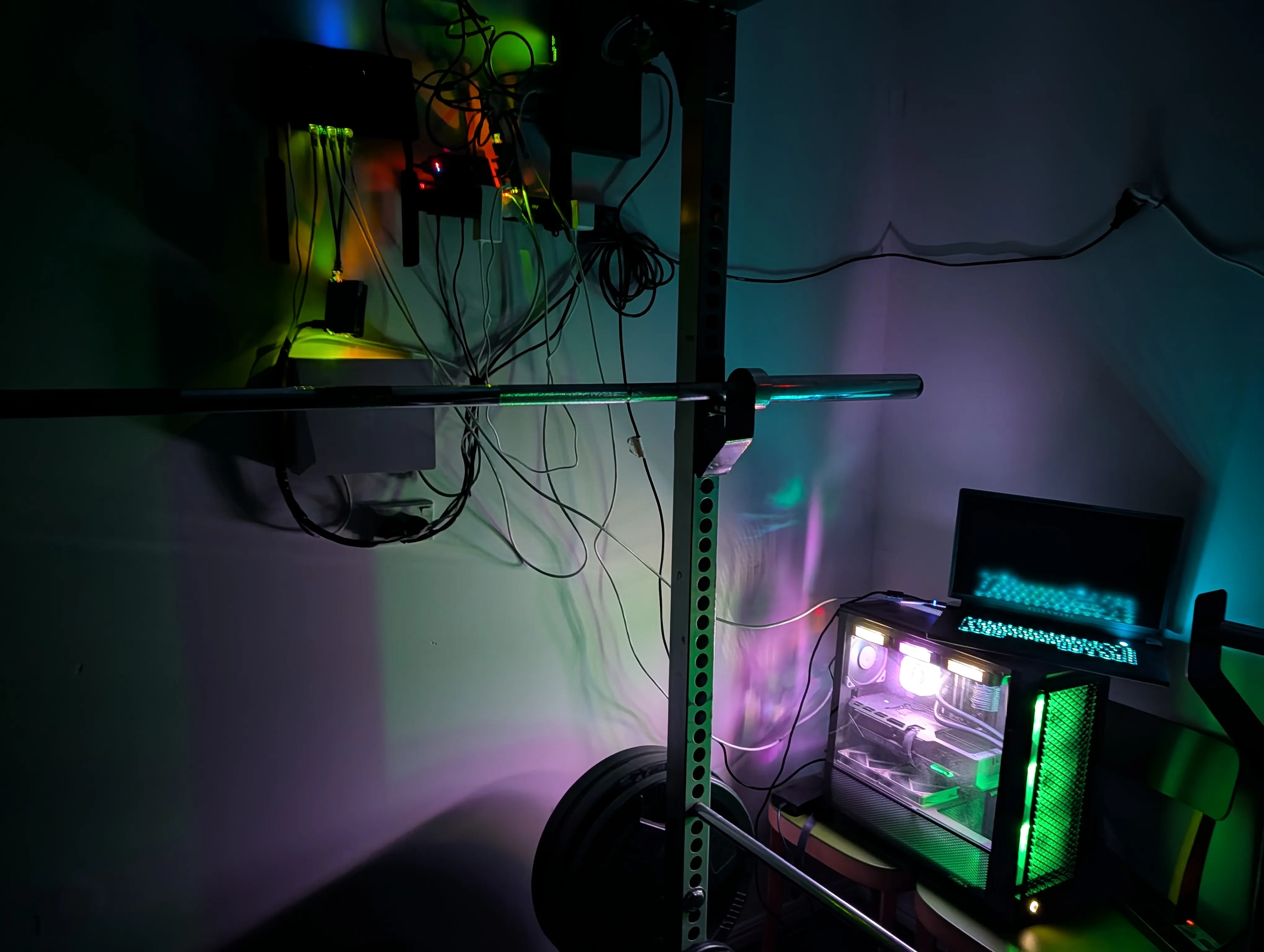 Home lab at night. A wall-mounted shelf of networking gear and single-board computers sits above a power rack, lit by RGB from a desktop machine beside it.